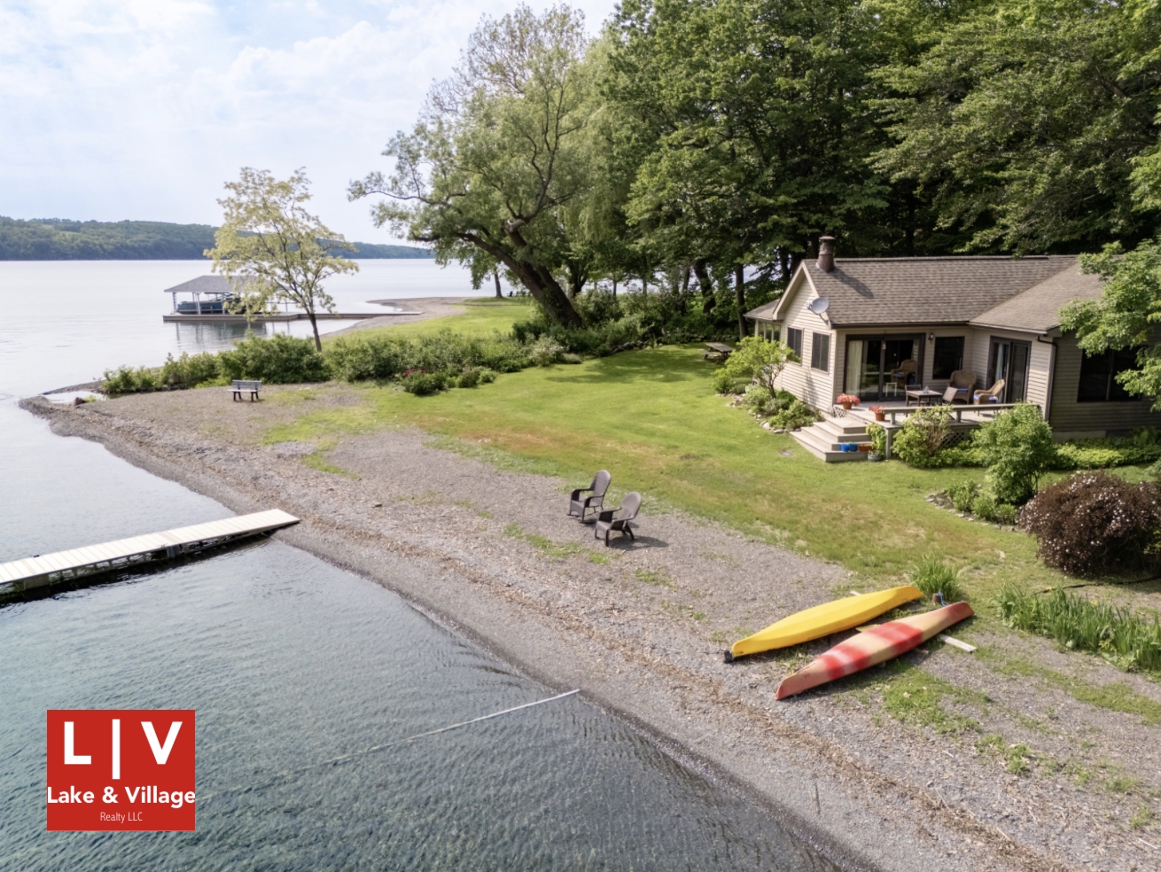 This image shows a lakeside cabin. It is on the east shore of Skaneateles Lake and on the viewer’s right. The cabin sits on a green lawn at the edge of the woods. The trees around the cottage are green. The lawn ends at a shale beach on which sit two grey outdoor chairs and two upside-down canoes. One canoe is red, & one is yellow. A dock runs from the beach on to the lake. The red-and-white Lake and Village logo is in the viewer’s lower left-hand corner.